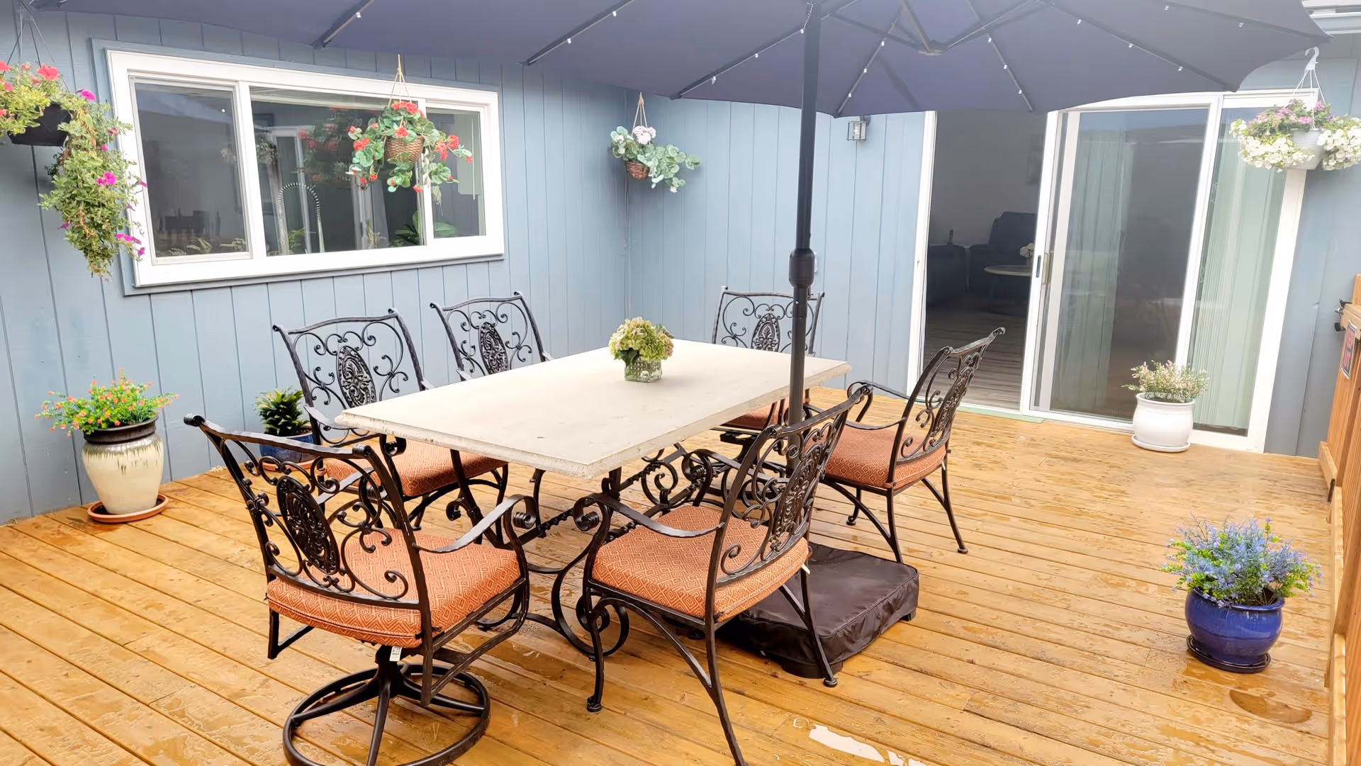 Outdoor patio area with a wooden deck featuring a rectangular table with six ornate metal chairs with orange cushions. A large umbrella is positioned in the center of the table. Several hanging flower baskets and potted plants decorate the space. Sliding glass doors lead inside the building with blue siding walls.