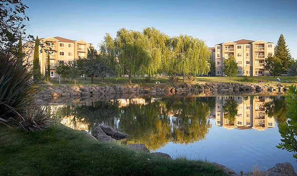 A serene outdoor scene at Westmont of Chico featuring a calm pond reflecting nearby trees and multi-story residential buildings under a clear sky.