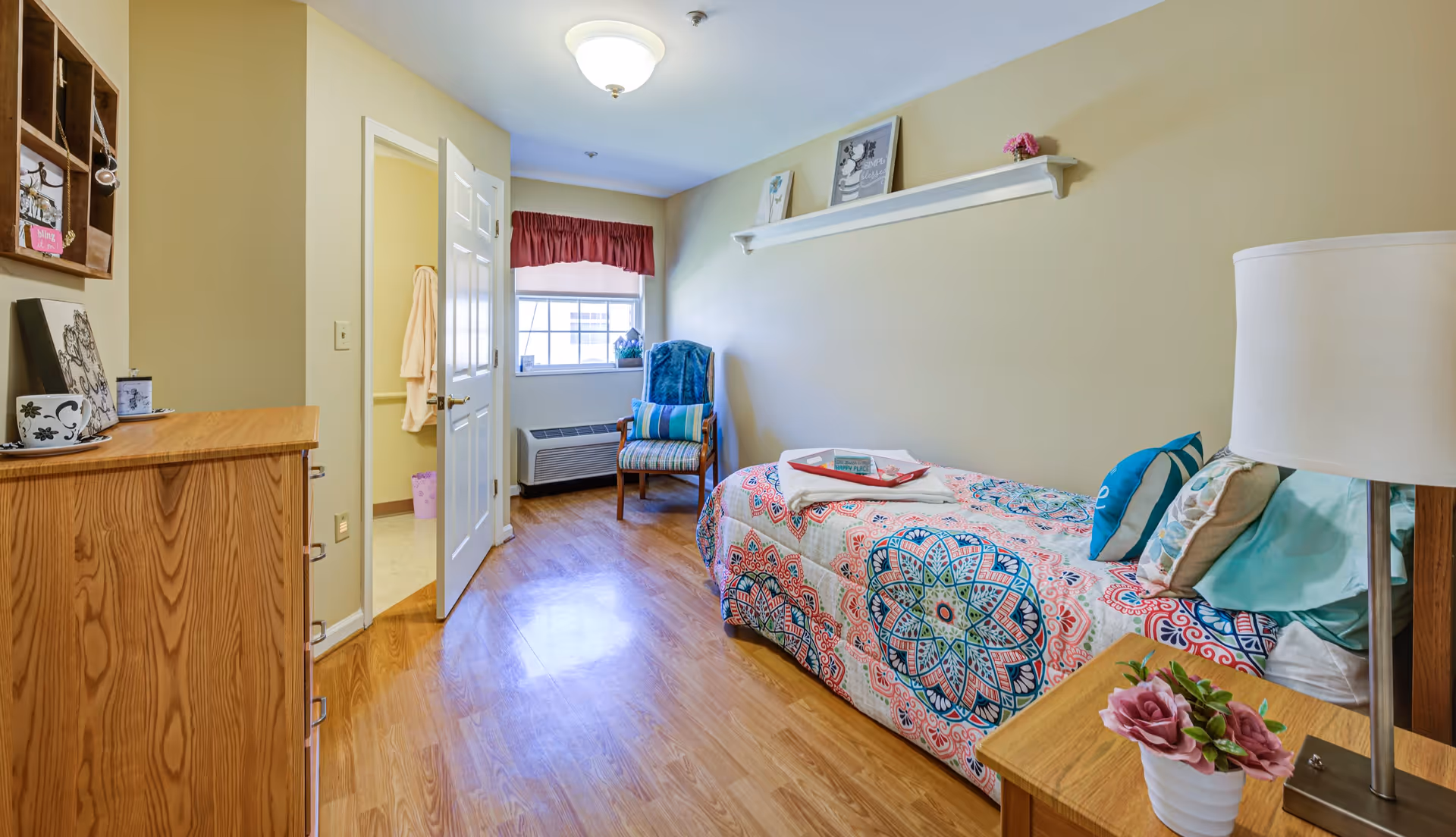 A cozy bedroom with a single bed covered in a colorful patterned quilt, a wooden nightstand with a lamp and a potted flower, a wooden dresser, a chair with a cushion near a window with a red valance, and a door leading to a bathroom.