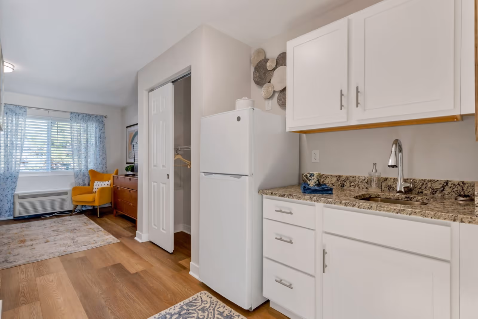 Interior view of a room in an assisted living facility featuring a small kitchen area with white cabinets, granite countertop, a sink with a modern faucet, and a white refrigerator. Adjacent to the kitchen is a cozy sitting area with a yellow armchair, a wooden dresser, a window with blue patterned curtains, and a closet with sliding doors.