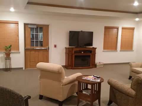 A cozy living room area with beige armchairs and a small wooden round table with magazines on it. A wooden cabinet with an electric fireplace and a flat-screen TV on top is against the wall. There are three windows with wooden blinds and a wooden door with a window. The room has recessed lighting and neutral-colored walls and carpet.