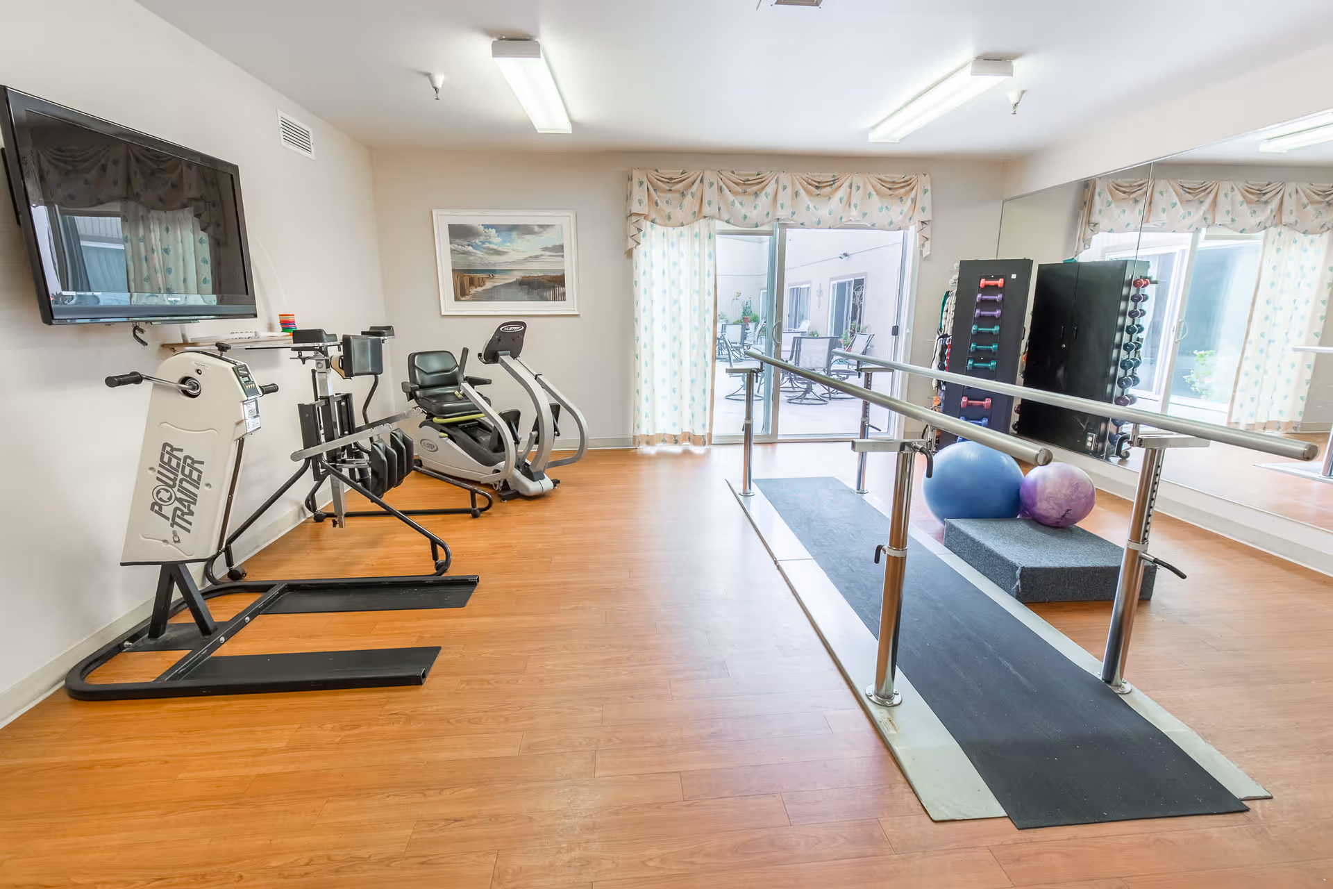 A rehabilitation or exercise room with wooden flooring, featuring parallel bars for walking practice, exercise balls, a rack of dumbbells, a recumbent exercise bike, and a Power Trainer machine. The room has large windows with patterned curtains, a wall-mounted TV, and a large mirror on one side reflecting the equipment and windows.