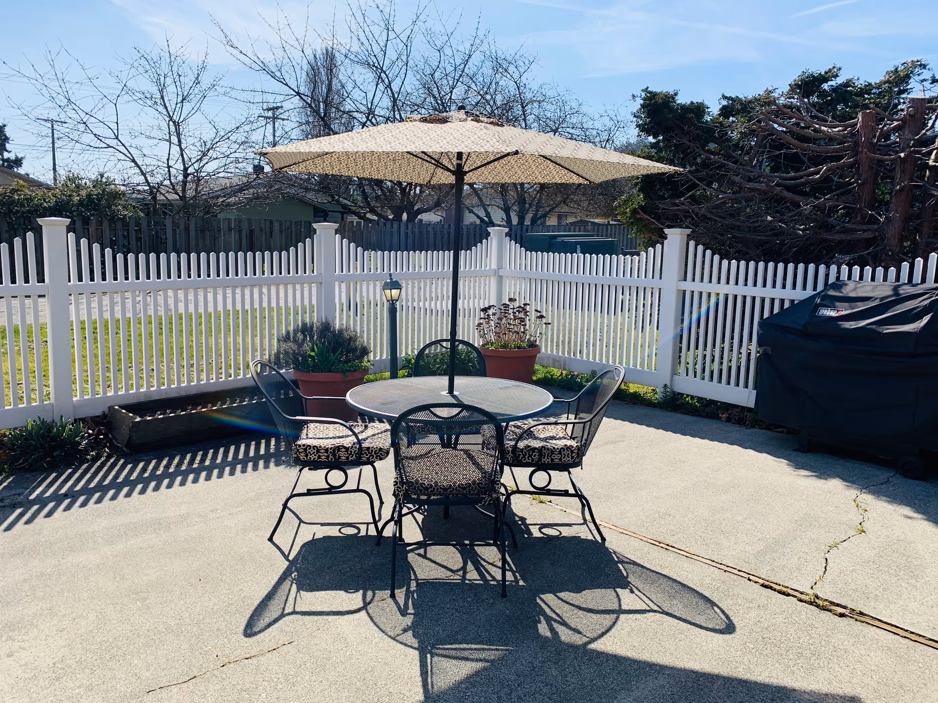 Outdoor patio area with a round metal table and four chairs with patterned cushions. A large beige umbrella is positioned over the table. The patio is enclosed by a white picket fence, and there are potted plants and a covered grill on the right side. Trees and a clear blue sky are visible in the background.
