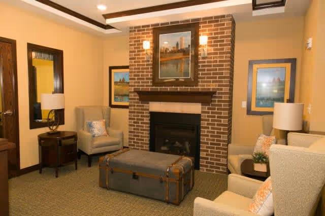 A cozy living room with a brick fireplace as the focal point. The room features two beige armchairs and a beige sofa with patterned cushions, a large rectangular ottoman in the center, two side tables with lamps, framed artwork on the walls, and warm yellow lighting.