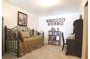 A cozy bedroom with a metal-framed daybed covered with a brown blanket and pillows. There is a framed picture hanging on the wall above the bed. Across from the bed is a wooden bookshelf filled with books and a desk with a chair. The walls are white and the floor is carpeted.