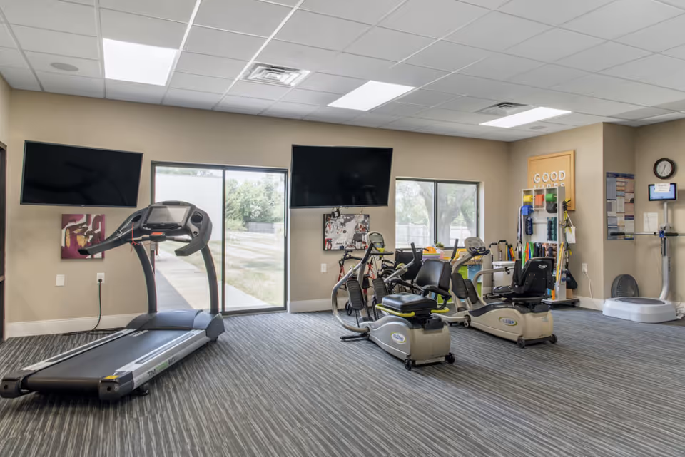 A senior living community exercise room with a treadmill, two recumbent exercise bikes, two wall-mounted flat screen TVs, large windows showing an outdoor view, and various exercise equipment and accessories organized on racks.