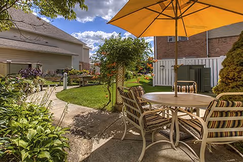 Outdoor patio area with a round table and four cushioned chairs under a large yellow umbrella. The patio is surrounded by greenery, including bushes and trees, with a paved walkway leading through the garden. Residential buildings are visible in the background under a partly cloudy sky.