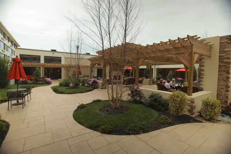 Outdoor courtyard area at The Addison of Cherry Hill featuring a paved walkway, landscaped garden beds with small trees and shrubs, wooden pergolas providing shaded seating areas, red umbrellas, and several elderly people sitting and conversing under the pergola.