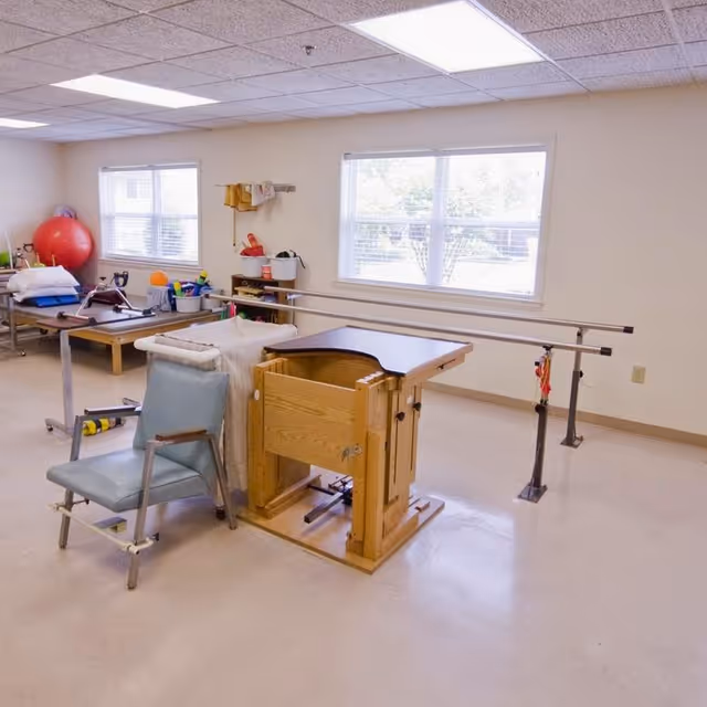A rehabilitation room with parallel bars for walking exercises, a padded chair with armrests, a wooden adjustable therapy table, and various therapy equipment including a large red exercise ball and other small items on a table near two windows letting in natural light.