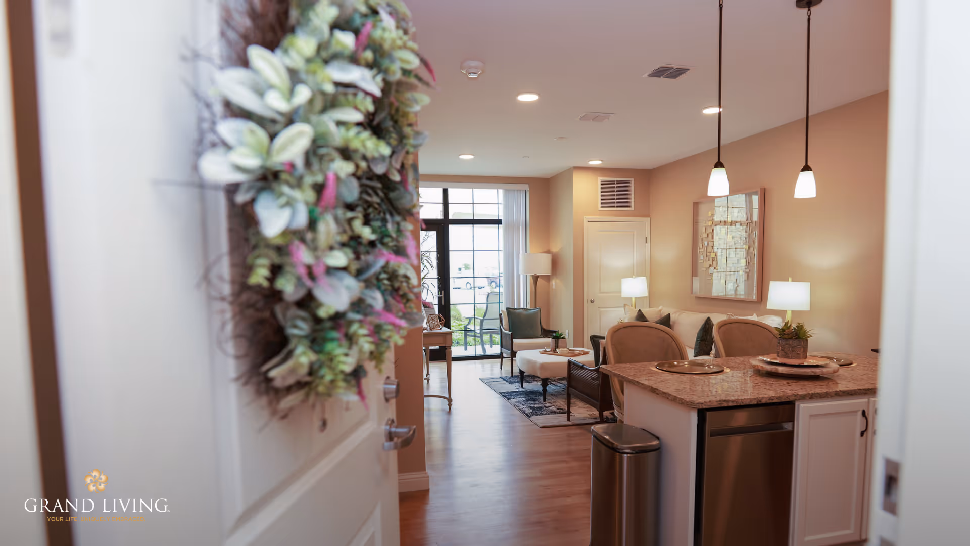 View through an open door decorated with a green and purple wreath into a warmly lit living space featuring a kitchen island with granite countertop, two pendant lights, a sitting area with chairs and a coffee table, and large windows letting in natural light.