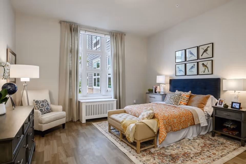 Well-lit furnished bedroom with a bed, nightstands, dresser, armchair, and a large window with curtains.