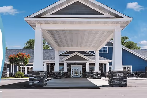 Front exterior view of a senior living facility with a covered entrance supported by white columns and stone bases. The building has blue siding, white trim, and a peaked roof. There is a tree with red flowers on the left side and a clear blue sky with some clouds in the background.