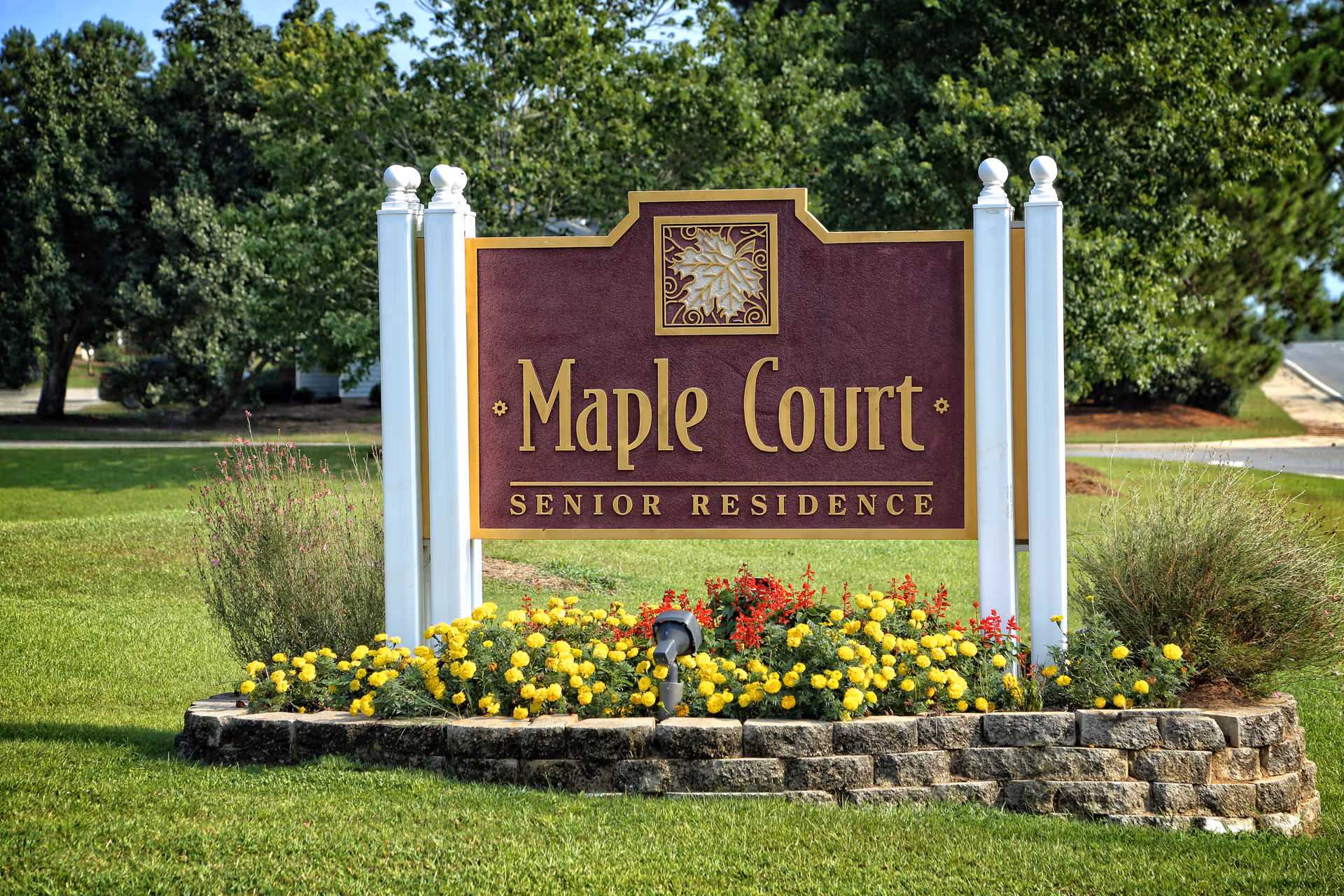 Entrance sign reading "Maple Court Senior Residence" surrounded by flowers and lawn.