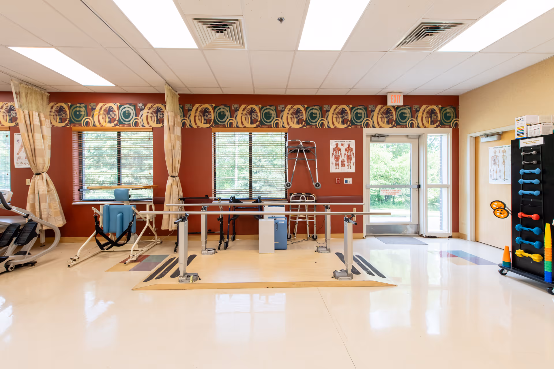 A physical therapy or rehabilitation room with parallel bars for walking exercises, various mobility aids such as walkers and a wheelchair, anatomical charts on the walls, and a rack with colorful exercise equipment. The room has large windows with blinds and patterned valances, beige and red walls, and a door leading outside.