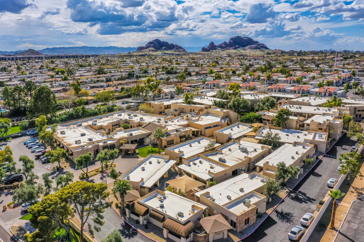 Aerial view of Scottsdale Village Square senior living facility with multiple beige buildings, parking areas, trees, and surrounding residential neighborhood under a partly cloudy sky with mountains in the background.