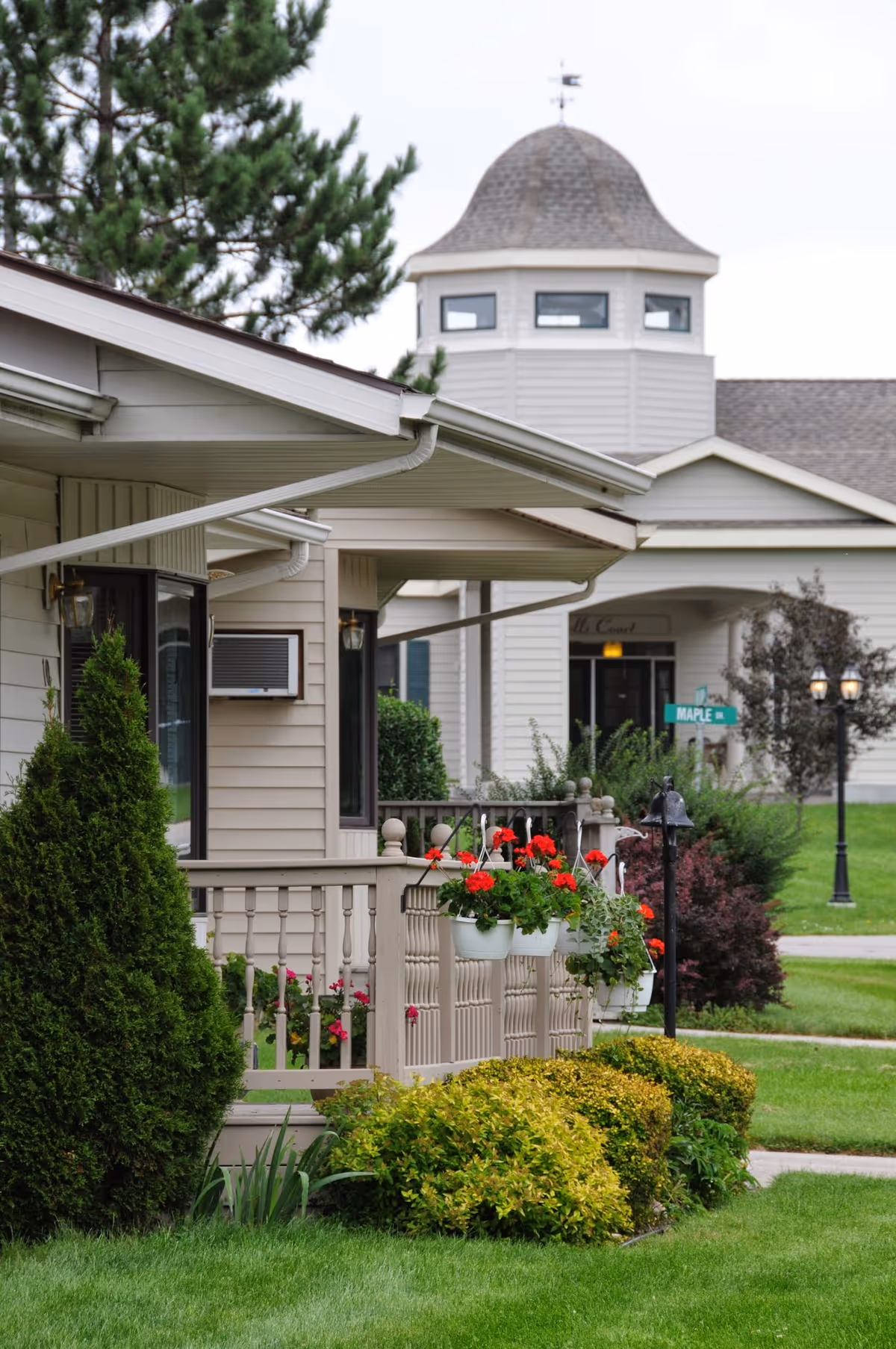 View of a residential building exterior with beige siding, a porch adorned with hanging flower pots containing red flowers, and well-maintained green bushes and lawn. In the background, there is a larger building with a distinctive dome-shaped roof and multiple windows, along with a street sign labeled 'MAPLE DR'.