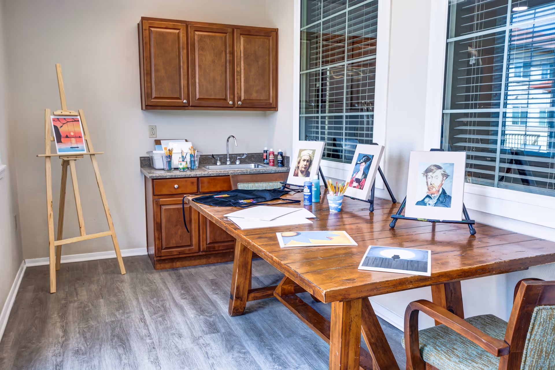 A bright art room with a wooden table displaying framed paintings and art supplies including paintbrushes and pencils. There is a wooden easel holding a colorful painting on the left side, and a countertop with a sink and wooden cabinets above and below it. The room has large windows with blinds and a wooden chair with armrests.