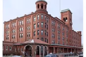 Exterior view of a large, historic brick building with multiple floors and arched windows, featuring two prominent towers on the roof. The building has a covered entrance and appears to be a senior living facility named Clifton Spring Spa Apartments.