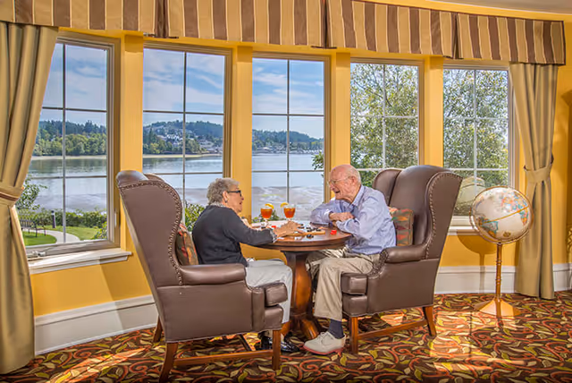Two elderly people sitting in large brown armchairs at a round wooden table playing a board game in a bright room with large windows overlooking a lake and trees. A globe on a stand is visible to the right, and the room has yellow walls and patterned carpet.