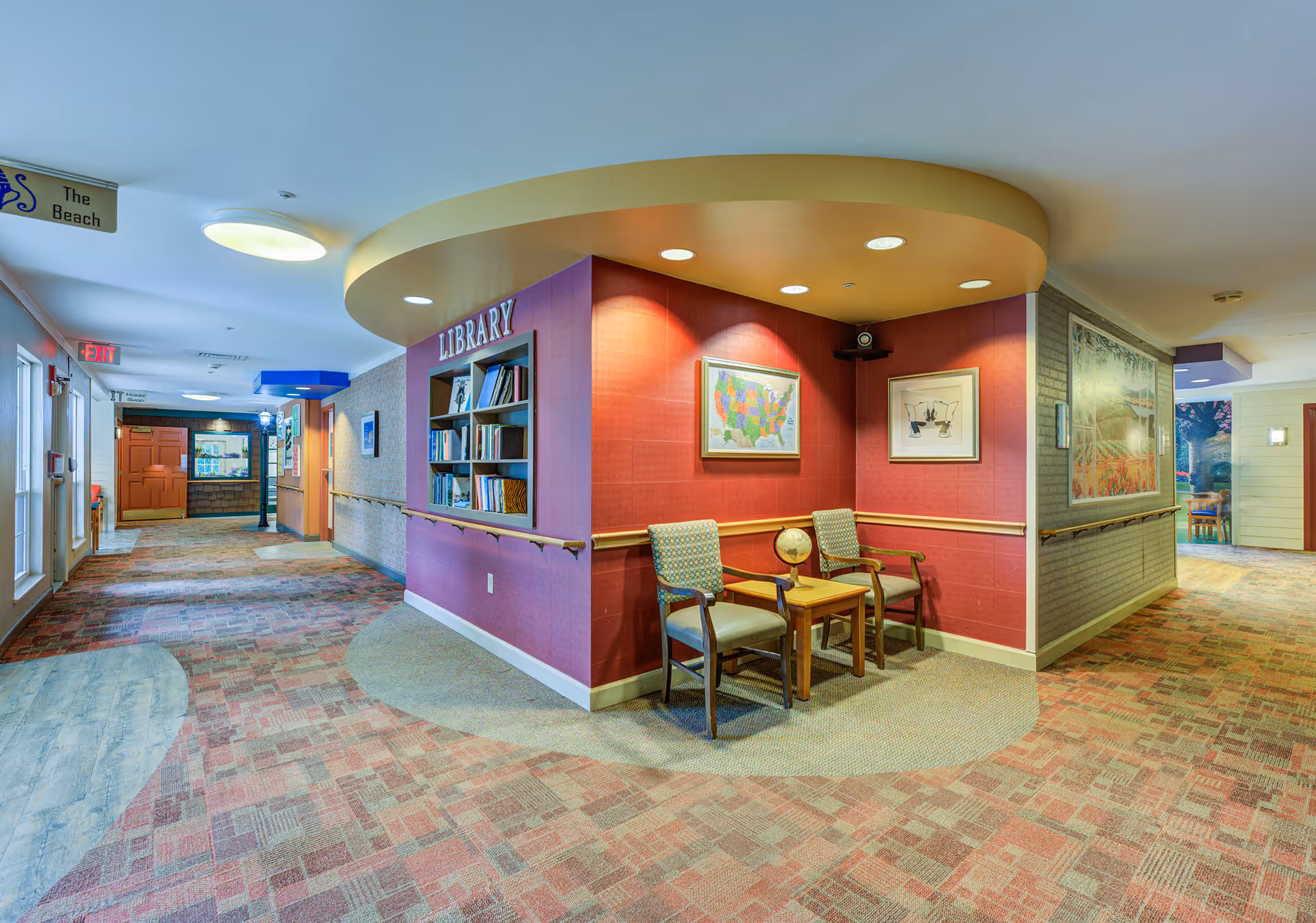 Interior hallway of Arden Courts - ProMedica Memory Care Community with a small library nook featuring bookshelves, two chairs, a small table with a globe, and framed artwork on the walls. The hallway has patterned carpet, handrails, and directional signs.