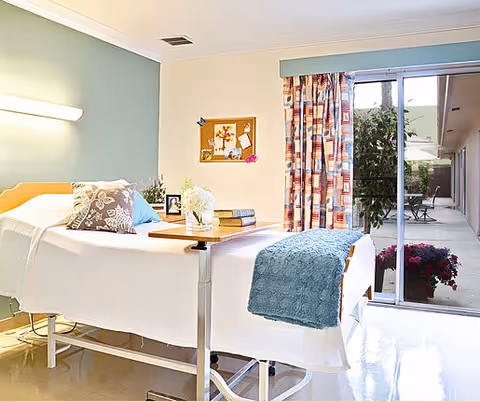 A bright and clean nursing center room with a hospital bed covered in white sheets and a blue blanket. There are decorative pillows on the bed, a small table with a vase of white flowers and books, a bulletin board on the wall, and a sliding glass door leading to an outdoor patio with plants and seating.