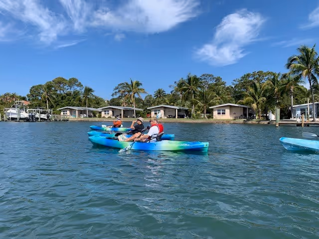 Two people kayaking on calm water near a shoreline with small houses and palm trees under a blue sky with some clouds.