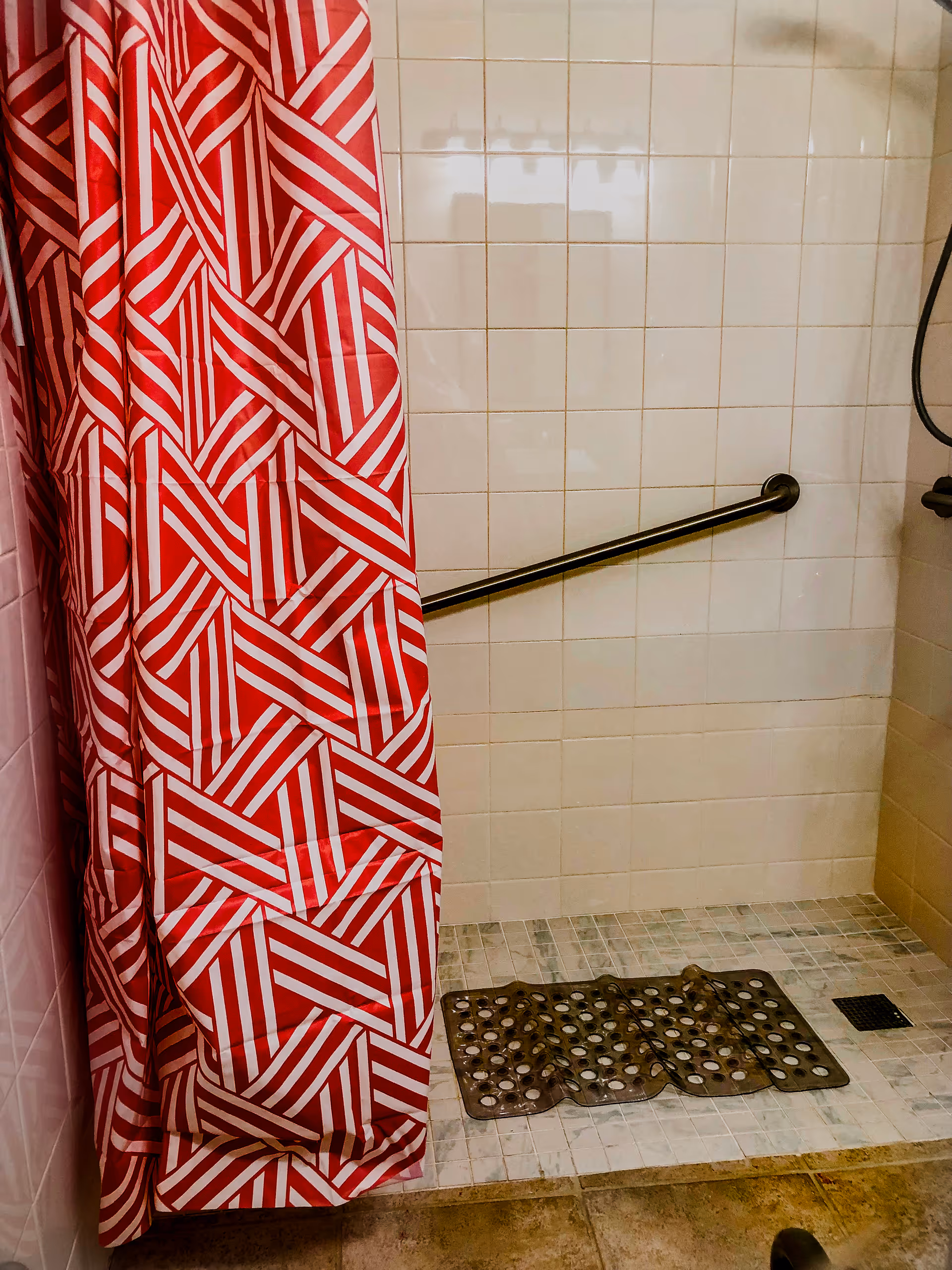 A tiled shower area with a red and white geometric patterned shower curtain partially drawn to the side. The shower has a metal grab bar mounted on the wall and a black non-slip mat on the floor.