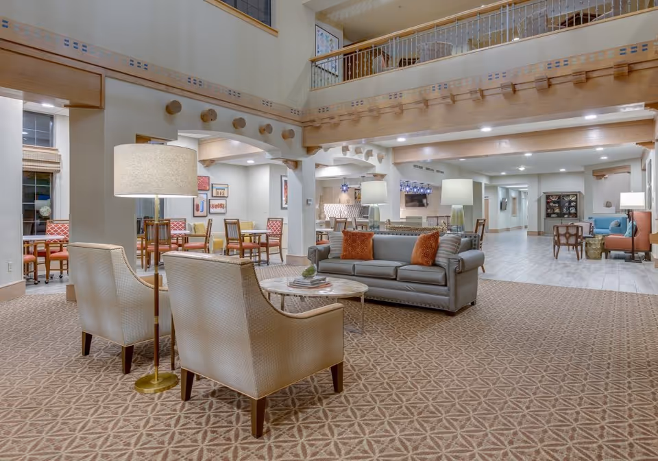 A spacious and well-lit senior living facility common area with a high ceiling and wooden beams. The room features a seating area with a gray sofa adorned with orange pillows, two beige armchairs, and a round marble coffee table. In the background, there are multiple tables and chairs arranged for dining or social activities, along with lamps and framed artwork on the walls. The flooring is a combination of patterned carpet and light-colored tiles.