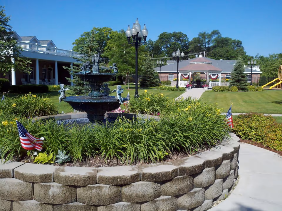 Outdoor garden area at Cottages of Fox Lake featuring a circular stone planter with green plants and a decorative fountain with cherub statues. American flags are placed around the planter. In the background, there is a walkway leading to a gazebo decorated with red, white, and blue bunting, surrounded by trees, lamp posts, and a playground slide on the right.
