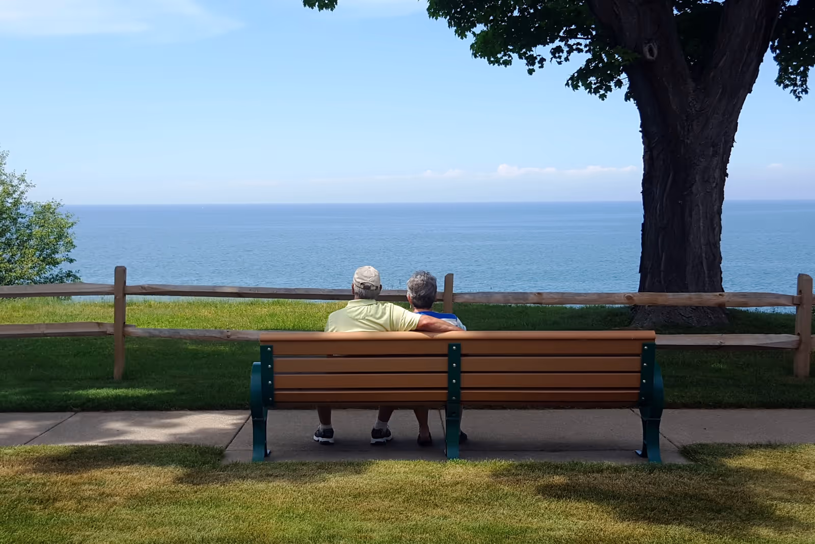 Two people sitting on a bench overlooking a large body of water with a tree and wooden fence.