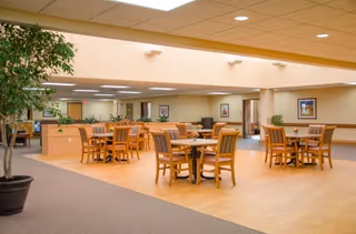 Spacious common area in a retirement community with multiple wooden tables and chairs arranged on a light wood floor. The room has beige walls, recessed lighting, and some potted plants, creating a warm and inviting atmosphere.