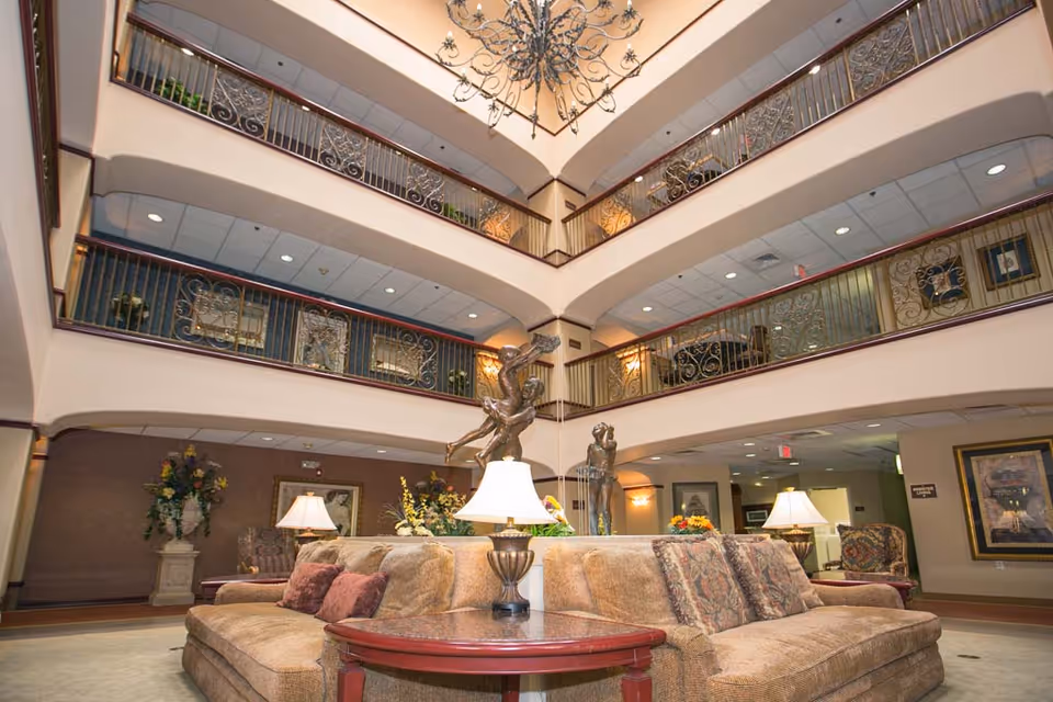 Spacious multi-level indoor common area with beige sofas arranged around a central table with a lamp and decorative sculptures. The area features ornate railings on the upper floors, a large chandelier hanging from the ceiling, and framed artwork on the walls.