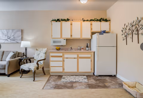 A small kitchenette area with white cabinets and a refrigerator, a microwave mounted above the counter, and a sink. To the left, there is a beige armchair with a floral pattern and a beige sofa with cushions. A floor lamp and a large floral wall art are also visible. On the right wall, there is a decorative metal wall art of trees and a pet bed on the floor.
