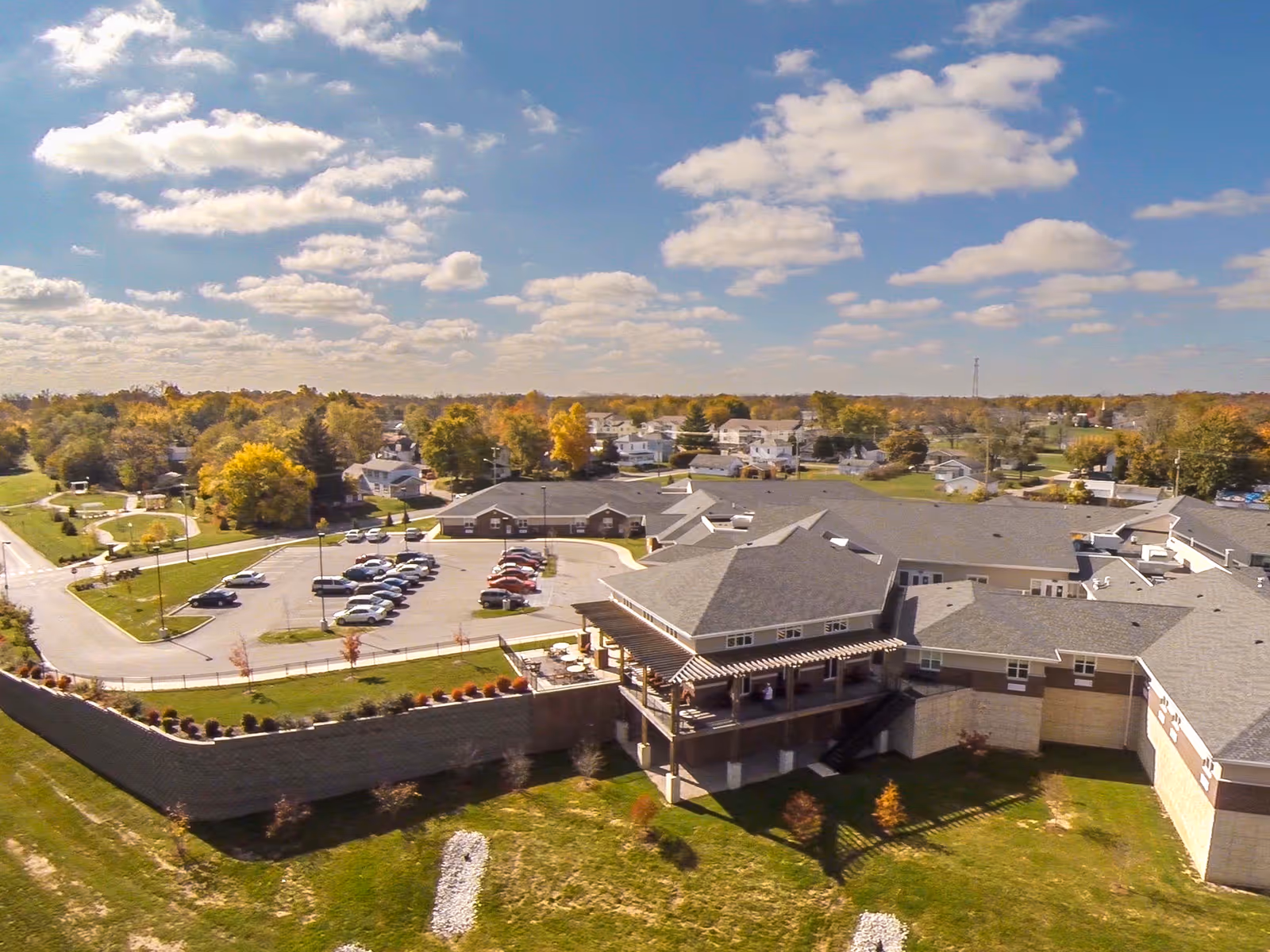 Aerial view of Aperion Care Marion facility showing a large building with multiple roof sections, a parking lot with several cars, surrounding green lawns, and a residential neighborhood in the background under a partly cloudy sky.