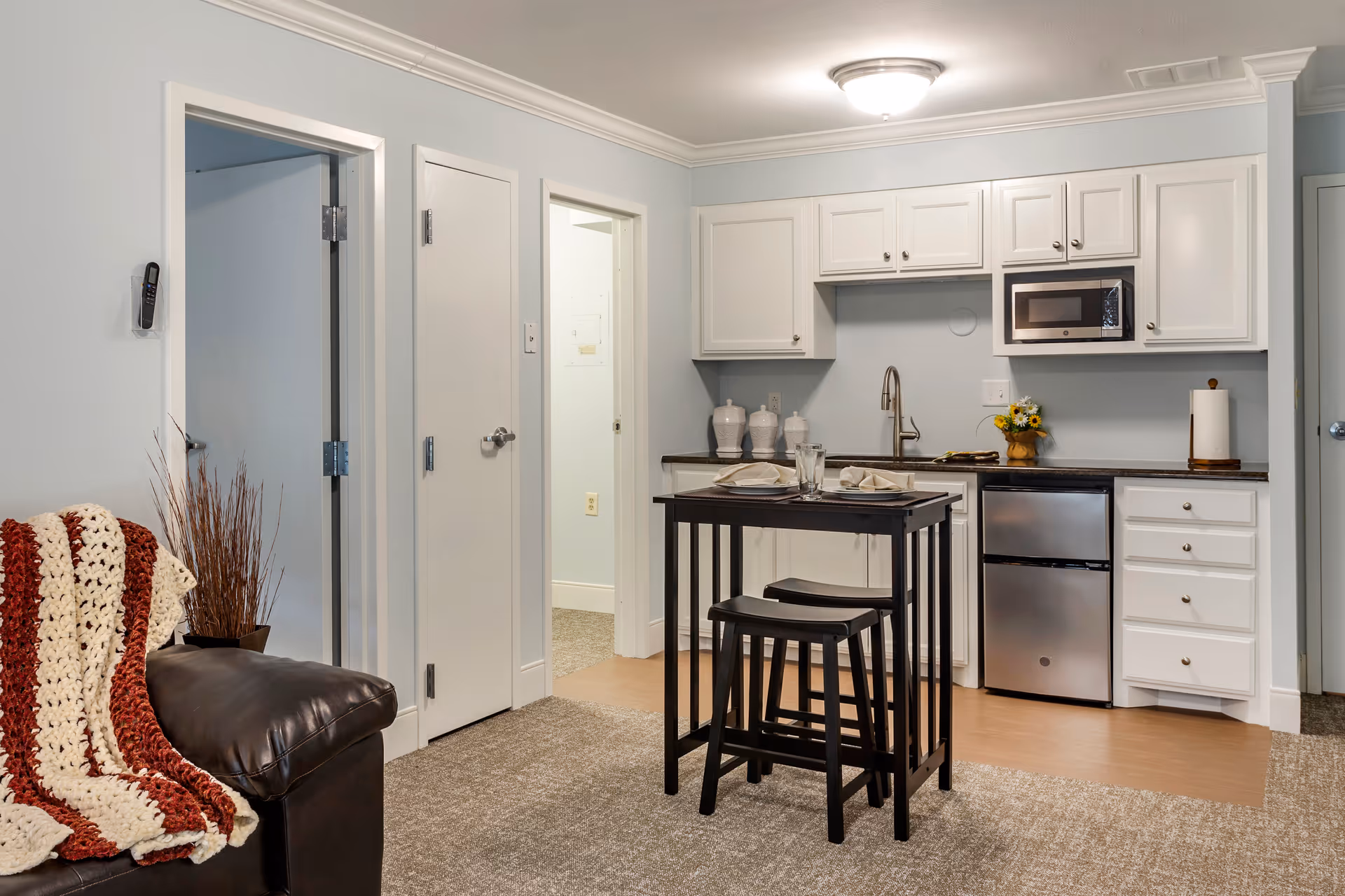 A small kitchen area with white cabinets, a stainless steel mini refrigerator, and a microwave. There is a black table with two stools set with plates and glasses in front of the kitchen. To the left, part of a dark brown leather couch with a red and white knitted blanket is visible. The floor is a combination of carpet and wood, and there are two closed doors and one open doorway in the background.