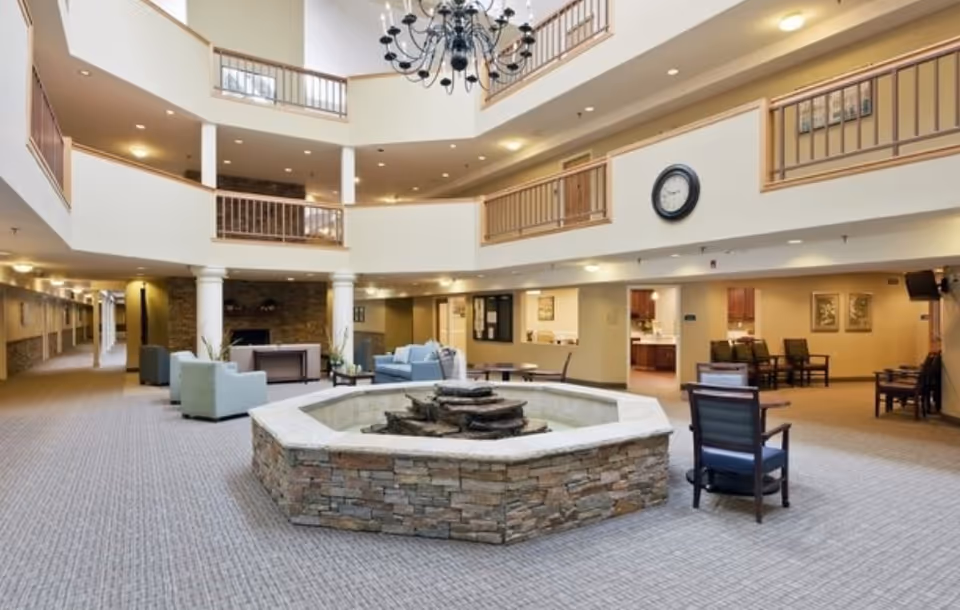 Spacious multi-level indoor common area with a stone water fountain in the center, surrounded by chairs and sofas. The area features beige walls, carpeted floors, a large black chandelier hanging from the ceiling, and a clock mounted on the wall. There are railings on the upper floors overlooking the space and a hallway extending to the left.