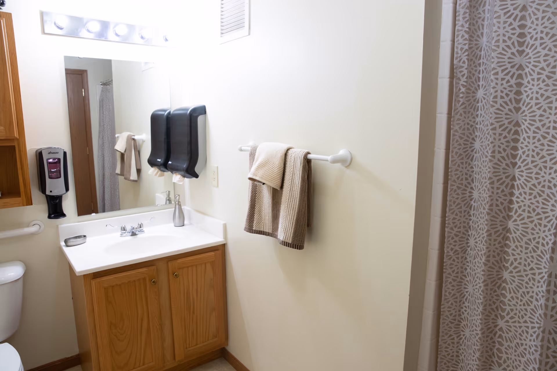 A bathroom with a wooden vanity cabinet, white countertop, and sink. Above the sink is a large mirror with a row of lights. Two black soap or paper towel dispensers are mounted on the wall next to the mirror. A towel rack on the adjacent wall holds two beige and brown towels. A patterned shower curtain is partially visible on the right side. A toilet is partially visible on the left side.