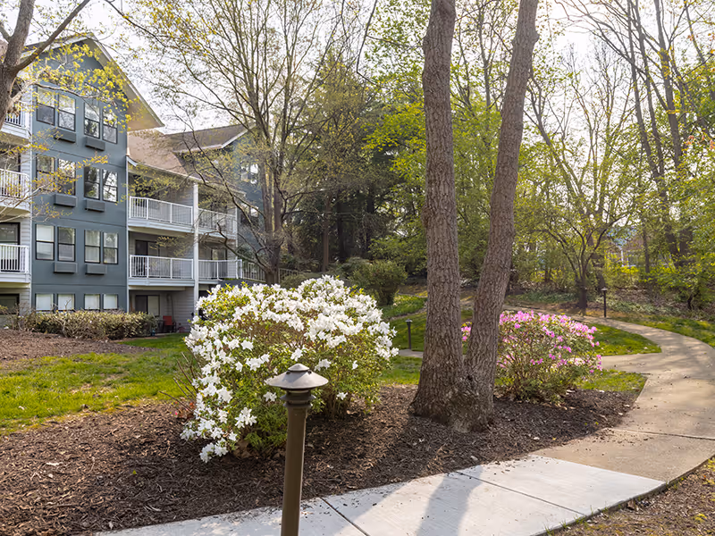 A paved walkway curves through a landscaped garden area with blooming white and pink flowers, trees, and shrubs. To the left, there is a multi-story residential building with balconies and large windows. The scene is bright and sunny, suggesting a peaceful outdoor environment.