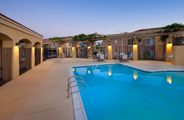 Outdoor swimming pool in a courtyard with pergolas, patio seating, and surrounding buildings at dusk.