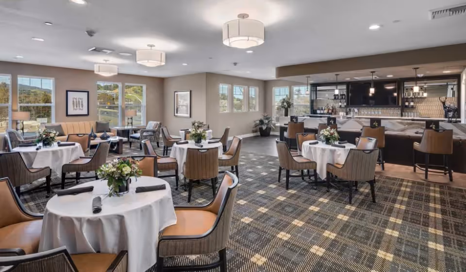 A spacious dining room with round tables covered in white tablecloths, each set with black napkins and floral centerpieces. The room features comfortable chairs, large windows letting in natural light, a patterned carpet, and a bar area with stools in the background.