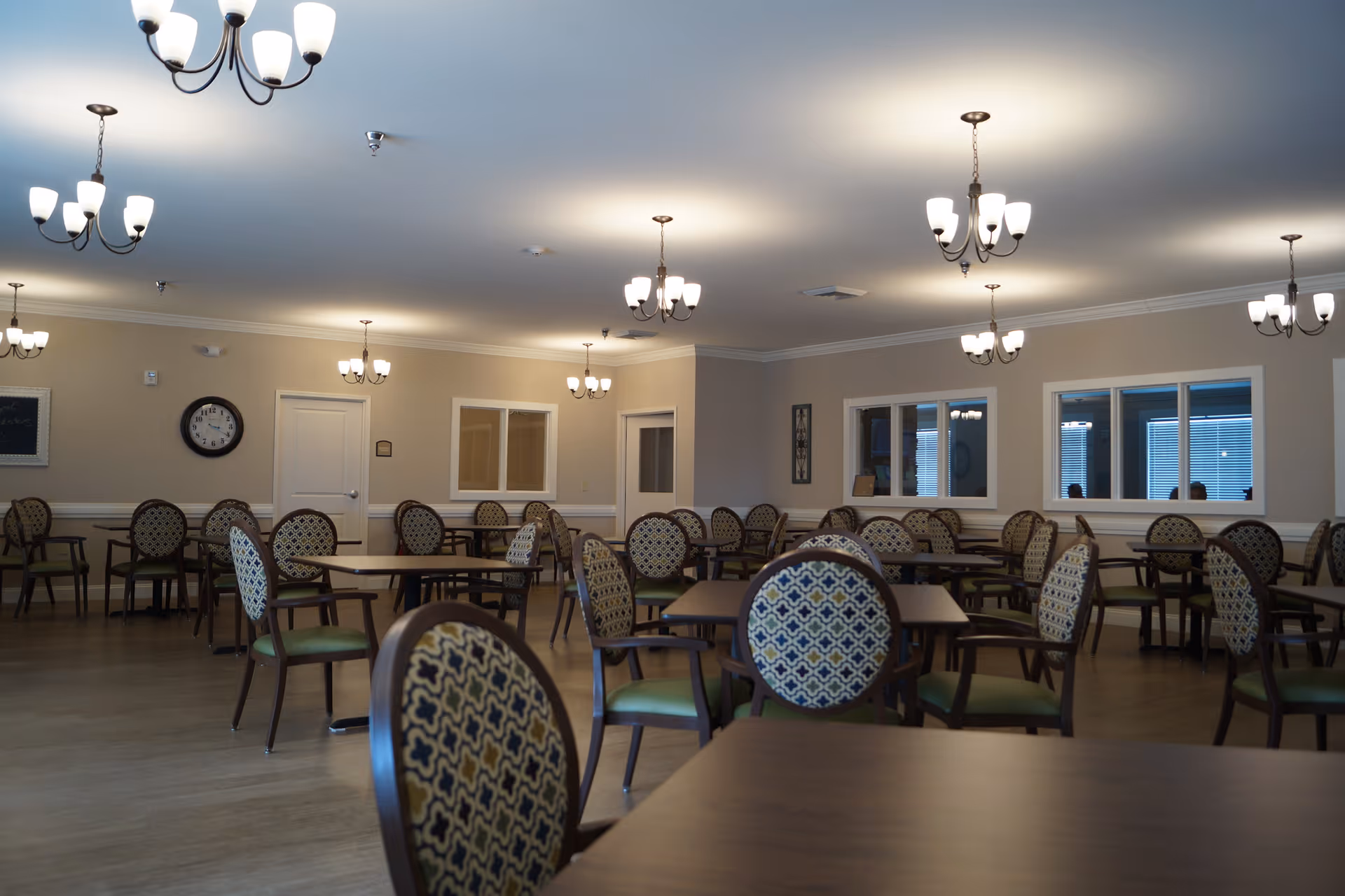 Large dining room with many round tables and upholstered patterned chairs lit by multiple chandeliers.