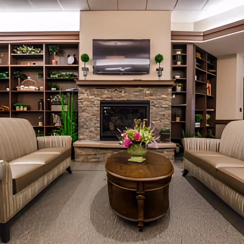 A cozy living room area with two beige upholstered sofas facing each other, a wooden oval coffee table with a floral arrangement in the center, a stone fireplace with a mounted flat-screen TV above it, and built-in wooden shelves filled with decorative items and greenery on either side of the fireplace.