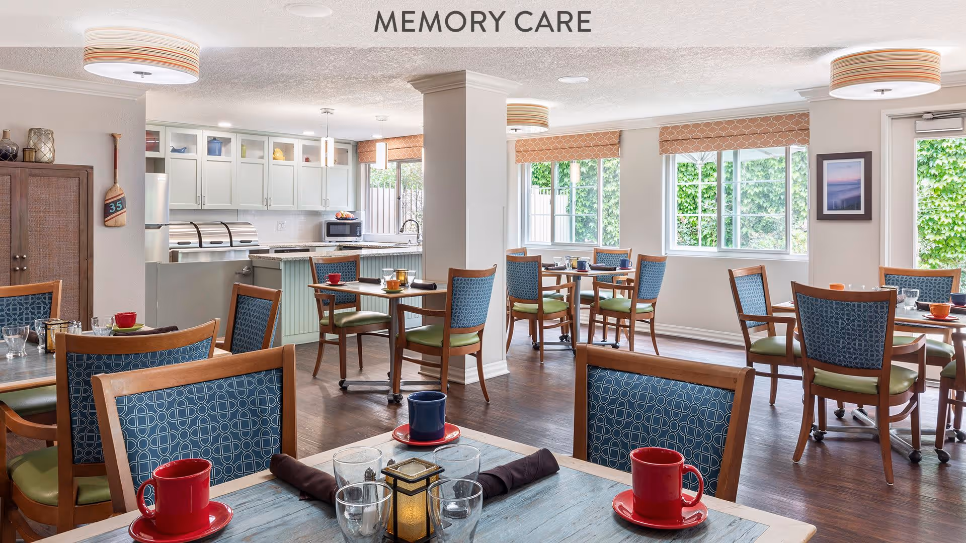 A bright and spacious dining area labeled Memory Care with multiple wooden tables and chairs featuring blue patterned backs and green seats. The room has large windows letting in natural light, a kitchen area with white cabinets and stainless steel appliances in the background, and modern ceiling lights. The floor is dark wood, and the walls are light-colored with some framed artwork and a door leading outside.