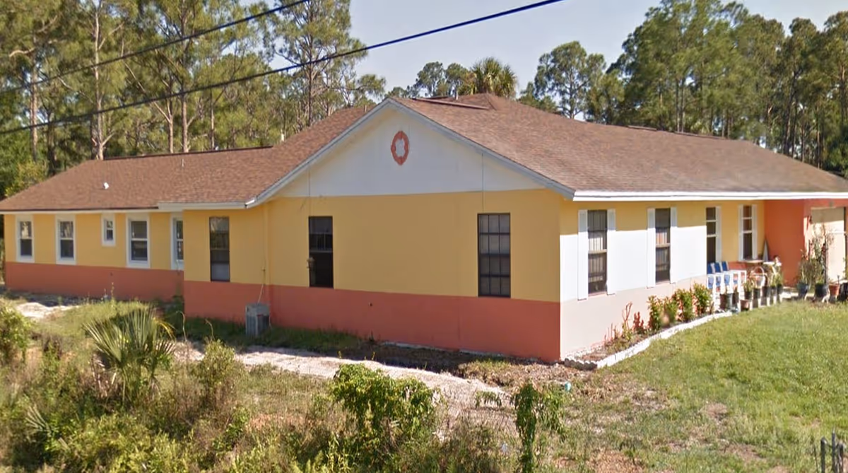 Exterior view of a single-story assisted living facility building with a brown roof, yellow and orange walls, several windows, and a small garden area with potted plants and greenery in front. The building is surrounded by trees and grass.