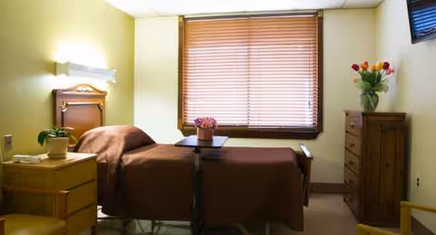 A single resident bedroom with a bed, dresser, window blinds, and flowers.