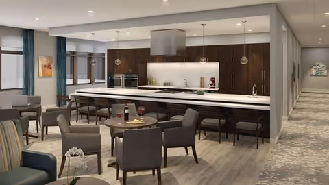 A modern senior living facility common area featuring a large kitchen with dark wood cabinets, a long white island with bar stools, and built-in ovens. The space includes several round tables with comfortable chairs, some with wine and snacks on them. Large windows with blue curtains allow natural light to fill the room, and a hallway with carpet leads off to the right.