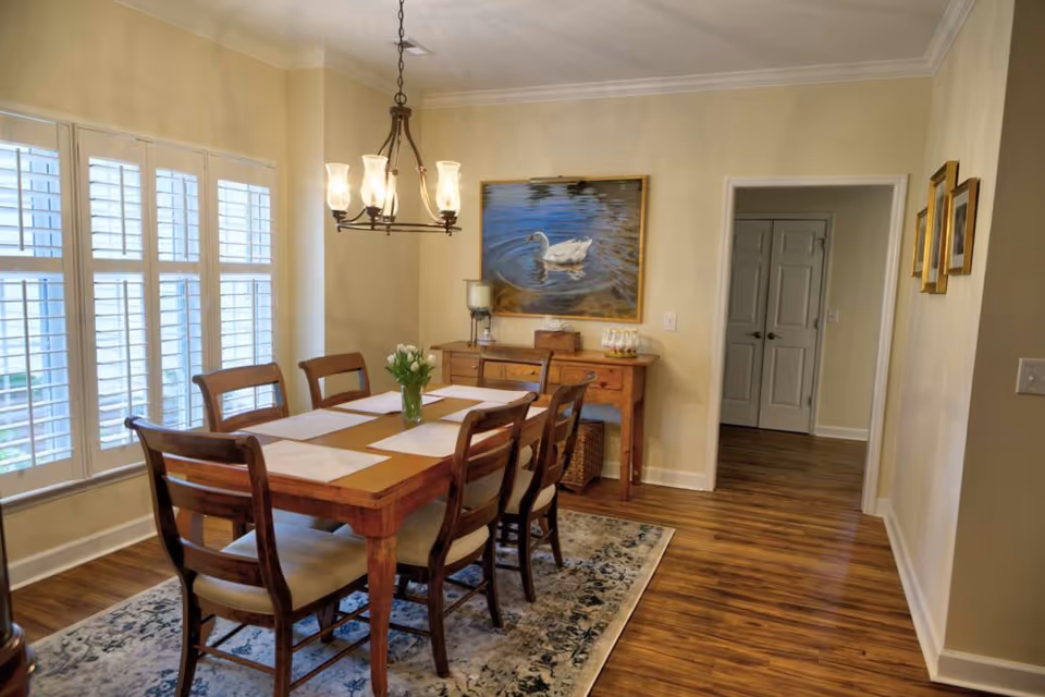 A bright dining room with a wooden table and six chairs under a chandelier, plantation-shuttered windows, a sideboard, and a swan painting on the wall.