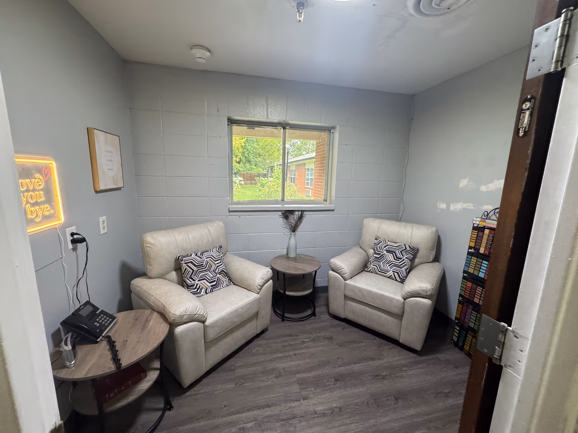 Small sitting room with two beige armchairs, side tables, a window, and decorative pillows.