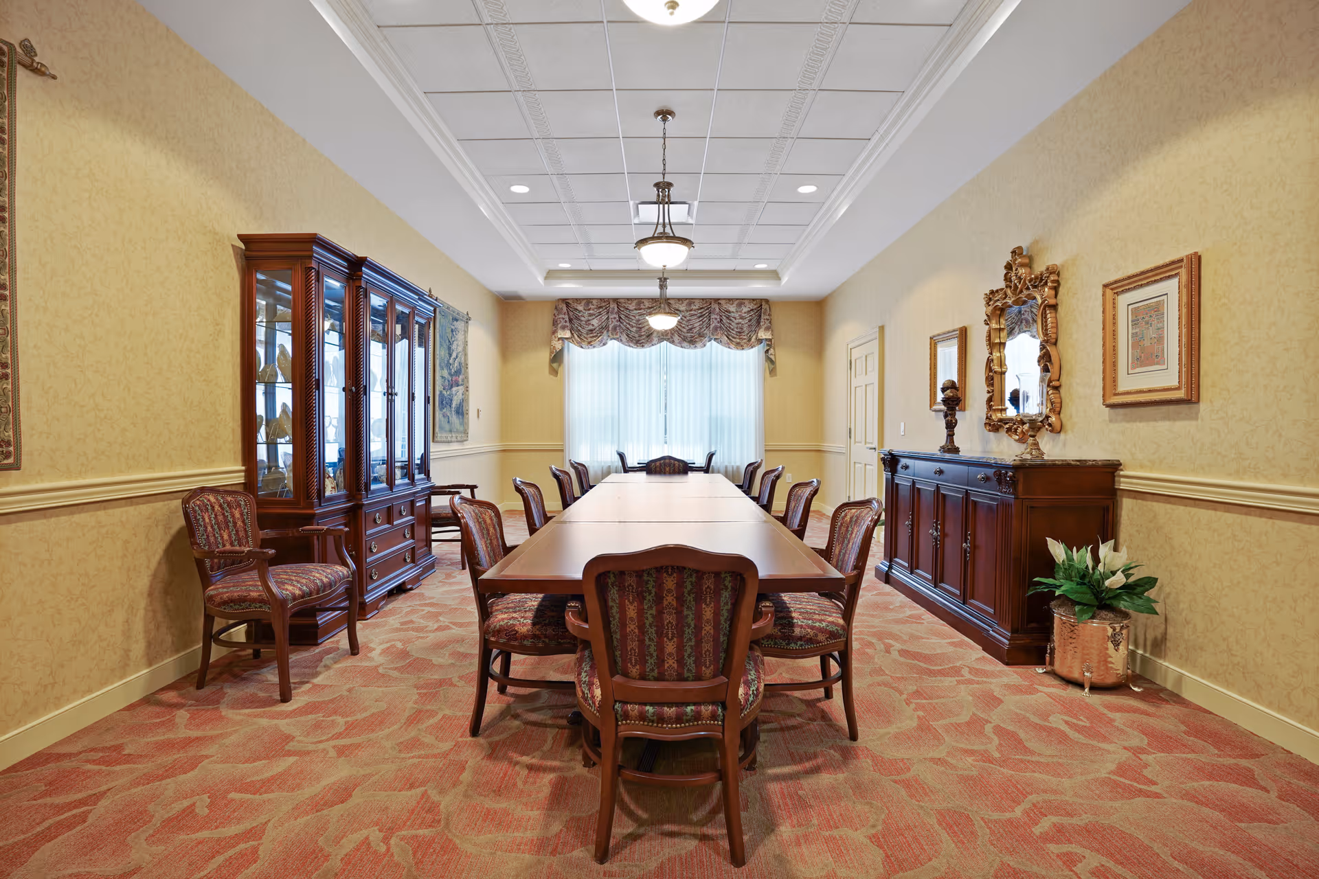 A formal dining room with a long wooden table surrounded by upholstered chairs. The room features patterned carpet, yellow wallpaper with a chair rail, a wooden china cabinet on the left, and a wooden sideboard with a decorative mirror and framed artwork on the right. A window with sheer curtains and a valance is at the far end of the room, and ceiling lights hang above the table.