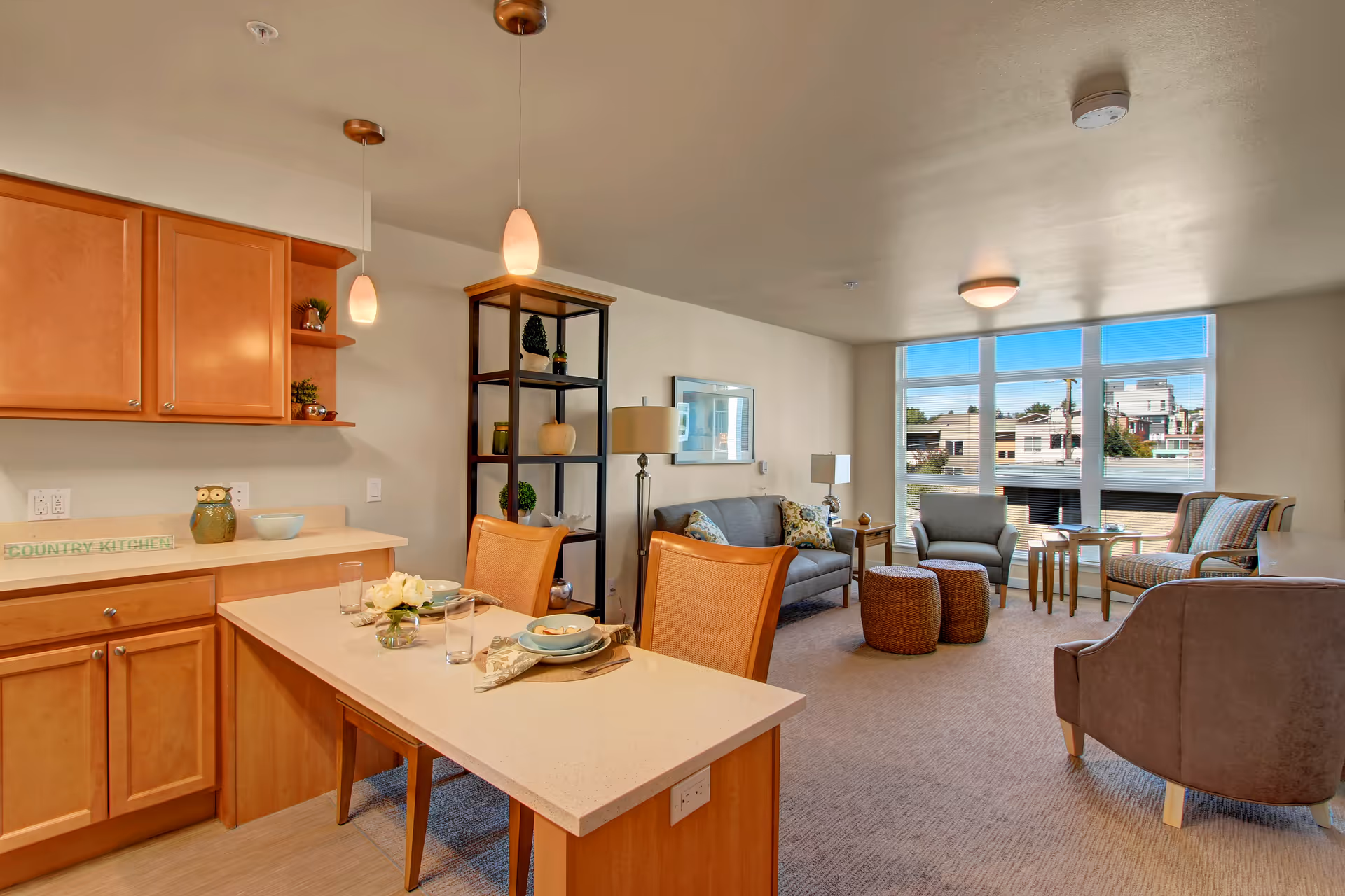 A bright and cozy living area in a senior living facility featuring a small kitchen with wooden cabinets and a countertop set for two. The living room has a gray sofa, two armchairs, two round woven ottomans, a floor lamp, and a shelving unit with decorative plants. Large windows let in natural light and offer a view of nearby buildings.